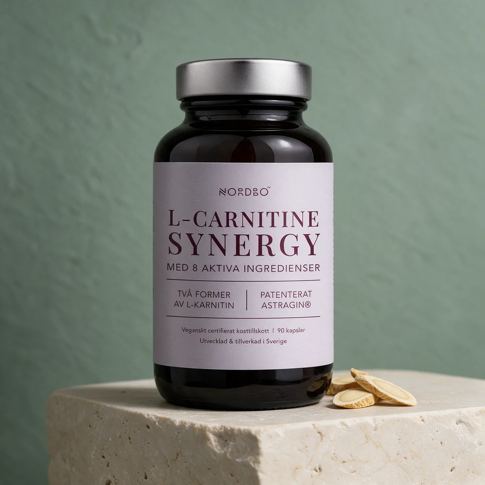 Nordbo L-Carnitine Synergy i glasskrukke med metallokk og «Med 8 aktiva ingredienser» på etiketten, ved skiver av astragalusrot