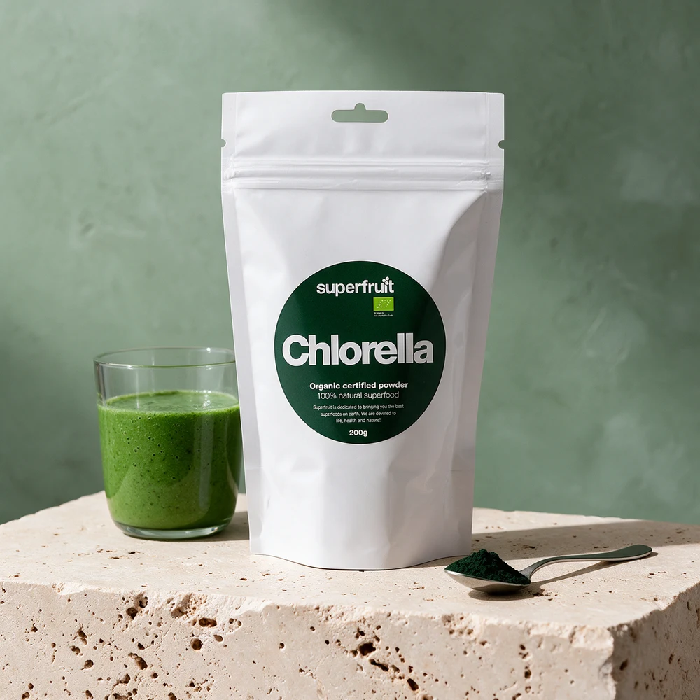 Superfruit Chlorellapulver Økologisk i en hvit pose med mørkegrønn rund etikett ved et glass grønn smoothie og en skje med grønt pulver på en travertinblokk
