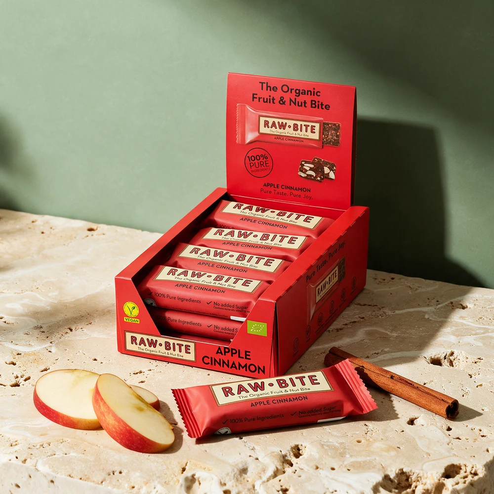 Rawbite Apple Cinnamon, åpen eske med tolv barer og én bar foran, med epleskiver og en kanelstang