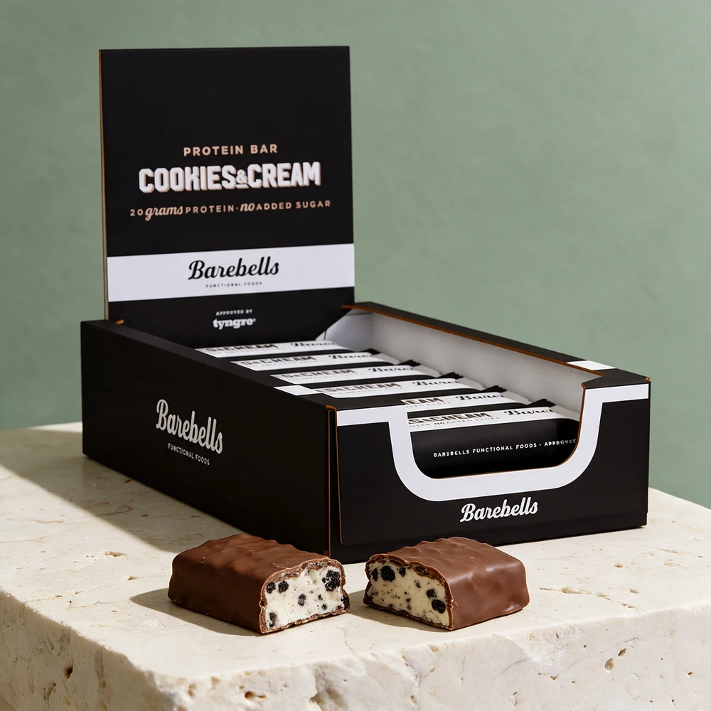 Barebells Protein Bar Cookies & Cream i eske og en bar med melkesjokoladetrekk delt i to, med kremhvit kjerne og mørke biter