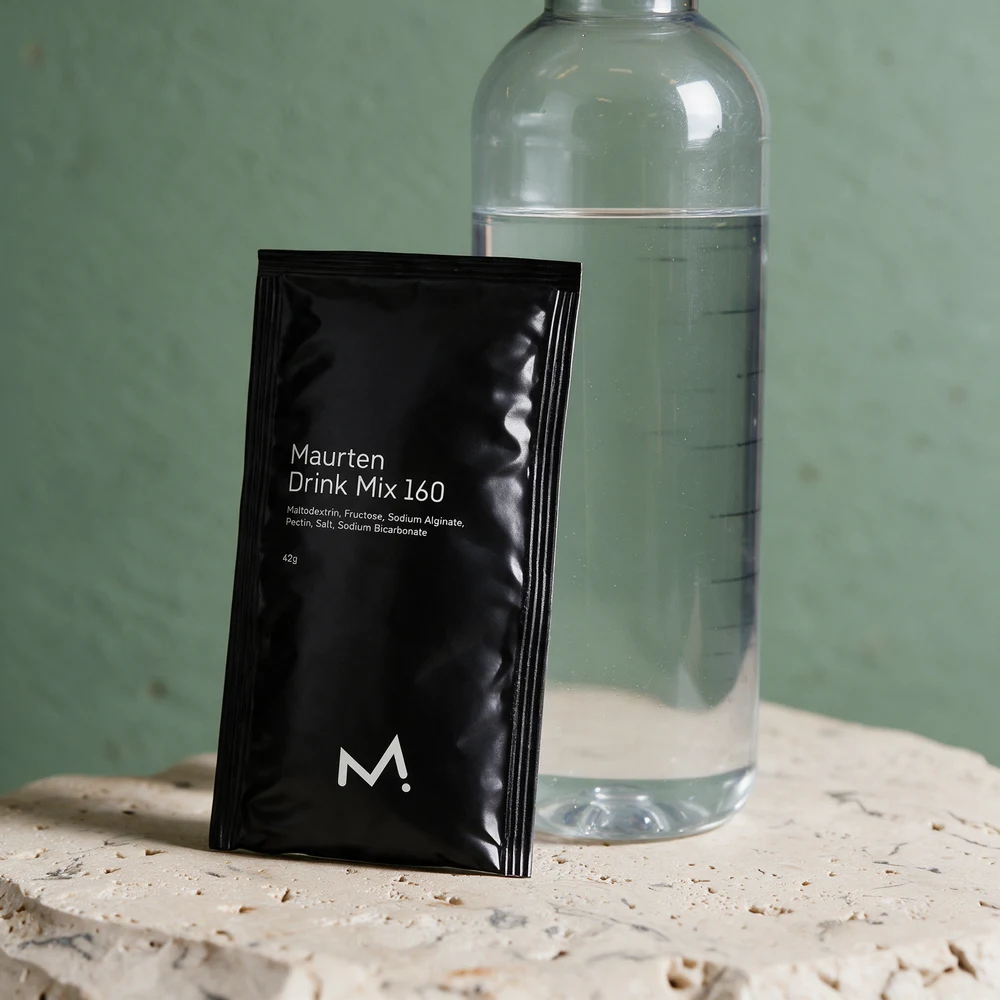 Maurten Drink Mix 160, svart porsjonspose, ved siden av en halvliter vann