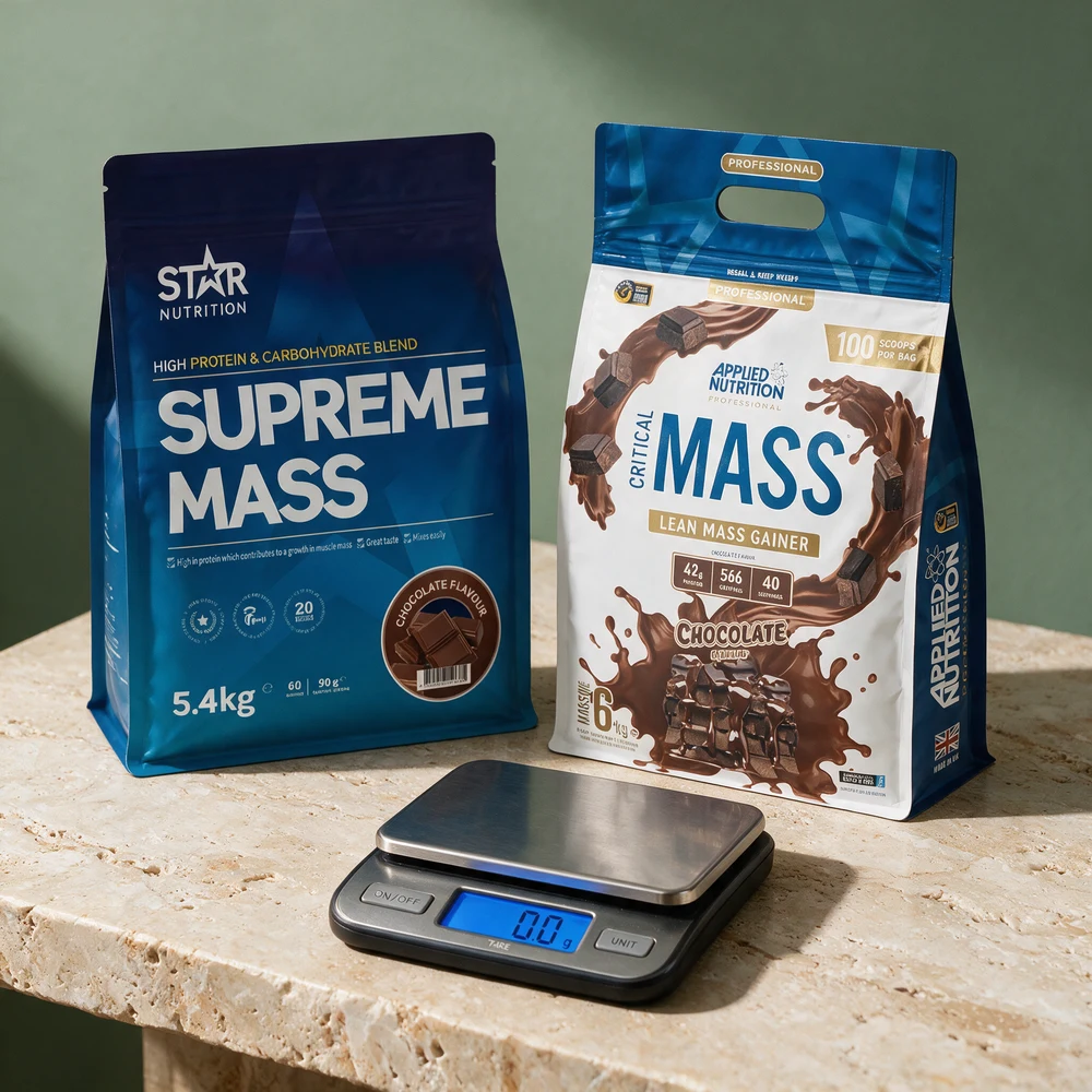Posene til Star Nutrition Supreme Mass og Applied Nutrition Critical Mass side om side med en sølvfarget digital kjøkkenvekt foran på et travertinbord
